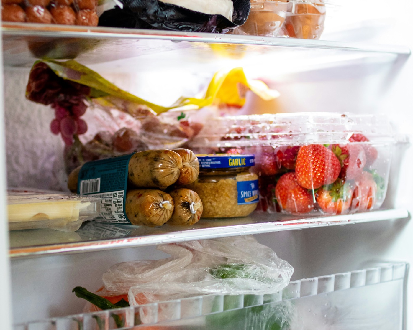 Open refrigerator interior — eggs, sausages, strawberries, garlic, and herbs across stocked shelves.
